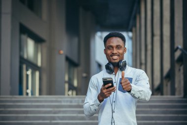 Kazanmaktan mutlu olan genç Afrikalı Amerikalı adam telefondaki sevinçlere ve günlük kıyafetlerdeki gülümsemelere bakıyor.