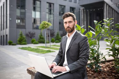Ciddi bir bankacı, laptoplar üzerinde çalışırken kameraya bakıyor. Kendine güvenen bir işadamı, ofisin yanında oturuyor.