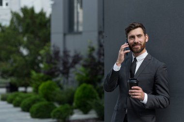 Başarılı iş adamı takım elbiseli bir simsar dışarıda ofisin yanında telefonla konuşuyor, bankacı öğle arasında elinde bir fincan kahve tutuyor gülümsüyor ve kameraya bakıyor.