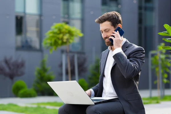 Successful male businessman reports good news by phone, working with laptop at lunchtime near office sitting on bench