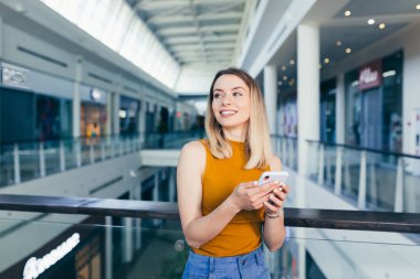 Ellerinde cep telefonu olan, günlük giysiler içinde mutlu bir genç kadın. Sarışın kız modern binalarda akıllı telefon alışverişi yapıyor. Kentsel arka plandaki kadın büyük salon alışveriş merkezi, ofis kullanımı