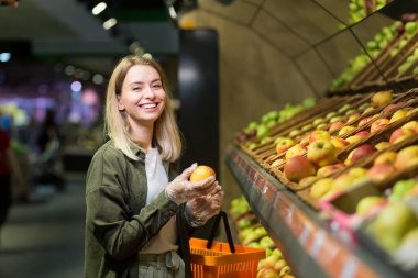 Genç sarışın kadın süpermarkette sebze topluyor. Markette alışveriş yapan kadın elinde sepetle sebze mağazasının yanında duruyor. Elmayı inceler
