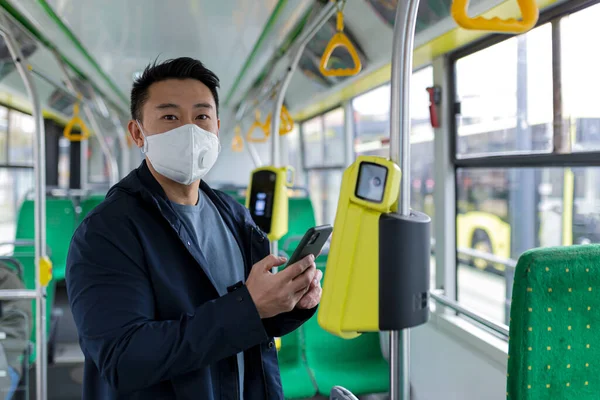Asian man in public transport bus in protective medical mask buys e-ticket by phone, passenger in empty bus