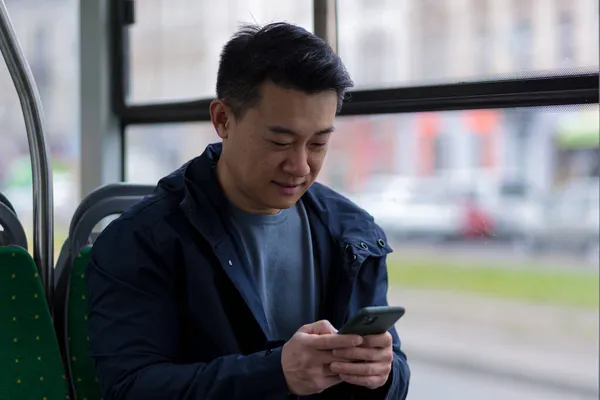 Young asian man corresponds and reads the news online, sitting in the bus, a passenger in casual clothes smiles