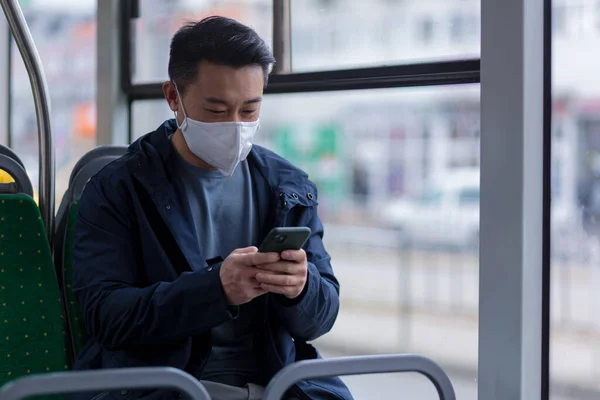 An Asian passenger in a protective medical mask on his face anxiously writes and reads news from a mobile phone, a man travels around the city by public transport bus