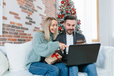 Genç bir aile babası ve karısı Noel hediyelerini online bir mağazadan seçiyorlar, evde dizüstü bilgisayarlarıyla oturuyorlar.