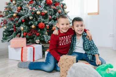Noel ağacının yanında oynayan iki küçük çocuk gülümseyip kameraya bakıyor.