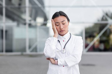 Klinik yakınında beyaz elbiseli genç Asyalı kadın, yorgun, depresif, yoğun çalışma gününden sonra depresyonda.