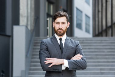 Genç kendinden emin beyaz sakallı iş adamı kameraya bakıyor. Modern ofis binasının kentsel arka planında duran ciddi bir iş adamı. Erkek.