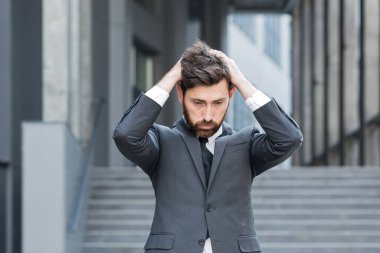 Üzücü bir iş adamı sakallı, beyaz bir adam kafasını tutuyor. İş adamı, girişimci, işçi ya da takım elbiseli bir işçi modern ofis merkezinin arka planında. Dışarı çıkın. Açık havada.