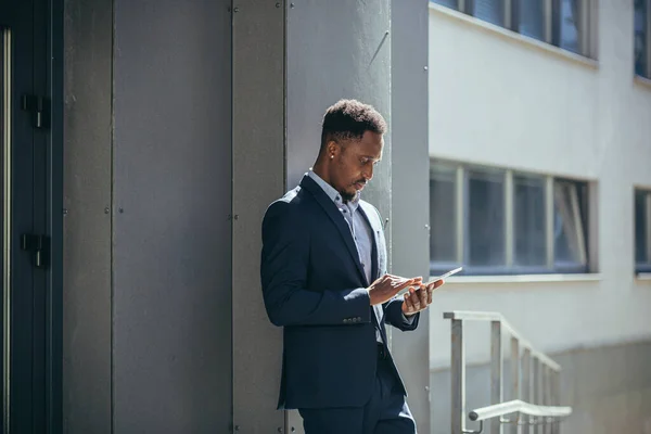 身穿正装的年轻的非洲裔美国商人手握平板电脑站在外面的现代化写字楼上工作 男子使用智能手机或手机室外城市街道 — 图库照片