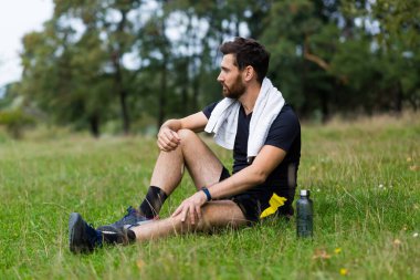 Antrenmandan yorgun düşmüş ya da dışarıda havluyla oturan bir sporcu. Portre terli fitness erkeği şehir parkında arka planda orman ve ağaçlarda. Kafkas sporcusu koştuktan sonra dinleniyor