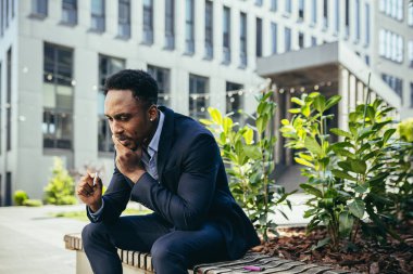 Afro-Amerikan iş adamı kenevir içiyor şehir parkındaki bankta oturuyor. Erkek işçi, iş adamı. Takım elbiseli bir ofis çalışanı dışarıda bir marihuanayı rahatlatıyor.