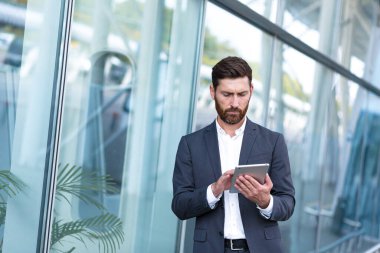 Resmi takım elbiseli şık sakallı iş adamı dışarıda arka plandaki modern ofis binasında elinde tabletle çalışıyor. Adam akıllı telefon kullanıyor ya da şehir dışında cep telefonu kullanıyor.