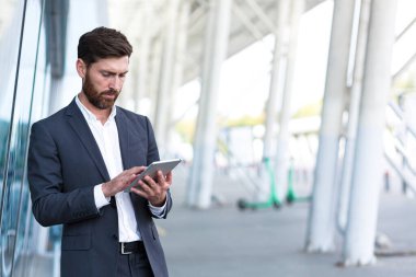 Resmi takım elbiseli şık sakallı iş adamı dışarıda arka plandaki modern ofis binasında elinde tabletle çalışıyor. Adam akıllı telefon kullanıyor ya da şehir dışında cep telefonu kullanıyor.
