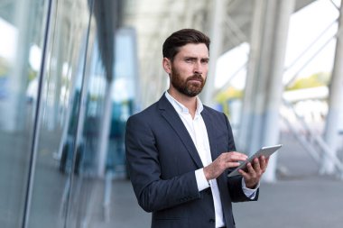 Resmi takım elbiseli şık sakallı iş adamı dışarıda arka plandaki modern ofis binasında elinde tabletle çalışıyor. Adam akıllı telefon kullanıyor ya da şehir dışında cep telefonu kullanıyor.