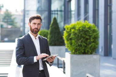 Resmi takım elbiseli şık sakallı iş adamı elinde tabletle dışarıda arka plandaki modern ofis binasında çalışıyor..