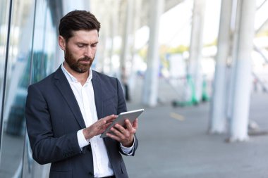 Resmi takım elbiseli şık sakallı iş adamı dışarıda arka plandaki modern ofis binasında elinde tabletle çalışıyor. Adam akıllı telefon kullanıyor ya da şehir dışında cep telefonu kullanıyor.