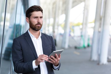 Resmi takım elbiseli şık sakallı iş adamı dışarıda arka plandaki modern ofis binasında elinde tabletle çalışıyor. Adam akıllı telefon kullanıyor ya da şehir dışında cep telefonu kullanıyor.
