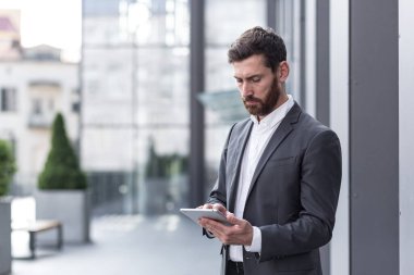 Resmi takım elbiseli şık sakallı iş adamı elinde tabletle dışarıda arka plandaki modern ofis binasında çalışıyor..
