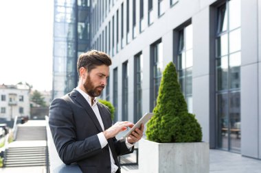 Resmi takım elbiseli şık sakallı iş adamı elinde tabletle dışarıda arka plandaki modern ofis binasında çalışıyor..