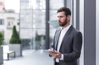 Resmi takım elbiseli şık sakallı iş adamı elinde tabletle dışarıda arka plandaki modern ofis binasında çalışıyor..