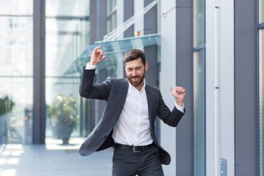 Neşeli mutlu iş adamı dans ederken şehrin arka planında modern bir ofis binası, açık havada komik takım elbiseli başarılı iş adamı zaferi kutluyor.