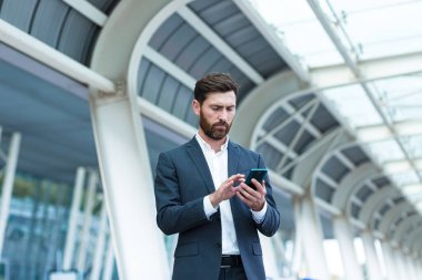 İş adamı arka planda duruyor modern bir tren istasyonu havaalanı ya da resmi takım elbiseli otobüs durağı uygulamalı cep telefonu kullanan seyahat eden akıllı telefonlu bir iş adamı bir taksi çağırır.