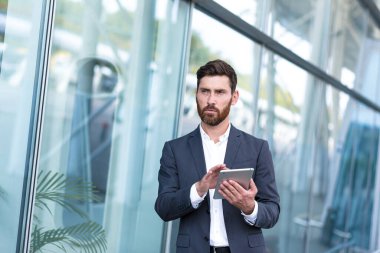 Resmi takım elbiseli şık sakallı iş adamı dışarıda arka plandaki modern ofis binasında elinde tabletle çalışıyor. Adam akıllı telefon kullanıyor ya da şehir dışında cep telefonu kullanıyor.