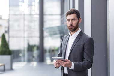 Resmi takım elbiseli şık sakallı iş adamı elinde tabletle dışarıda arka plandaki modern ofis binasında çalışıyor..
