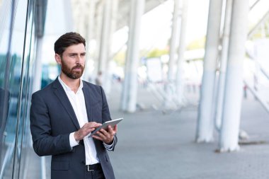Resmi takım elbiseli şık sakallı iş adamı dışarıda arka plandaki modern ofis binasında elinde tabletle çalışıyor. Adam akıllı telefon kullanıyor ya da şehir dışında cep telefonu kullanıyor.