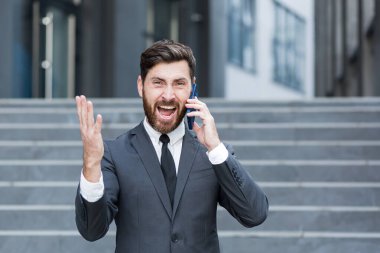 Kızgın erkek işadamı dışarıda telefonda konuşurken ağlıyor. Dışarıdaki çağrıda bağırıyor. Akıllı telefondan sinirli, profesyonel bir konuşma. Sinirli çalışan çığlığı Kentsel caddede konuşuyor.