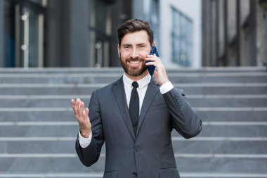 Başarılı genç iş adamı arka planda şehir merkezindeki modern ofis binasından bahsediyor. Sakallı iş adamı resmi takım elbise giymiş bir cep telefonu açık şehir caddesinde duruyor.