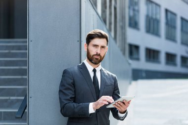 Resmi takım elbiseli şık sakallı iş adamı dışarıda arka plandaki modern ofis binasında elinde tabletle çalışıyor. Adam akıllı telefon kullanıyor ya da şehir dışında cep telefonu kullanıyor.