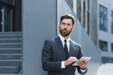Resmi takım elbiseli şık sakallı iş adamı dışarıda arka plandaki modern ofis binasında elinde tabletle çalışıyor. Adam akıllı telefon kullanıyor ya da şehir dışında cep telefonu kullanıyor.