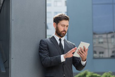Resmi takım elbiseli şık sakallı iş adamı dışarıda arka plandaki modern ofis binasında elinde tabletle çalışıyor. Adam akıllı telefon kullanıyor ya da şehir dışında cep telefonu kullanıyor.