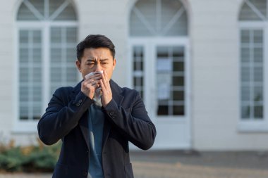 İş adamı yorgun ve hasta Asyalı adam sokaklarda hapşırıyor ve öksürüyor, alerjik reaksiyon.