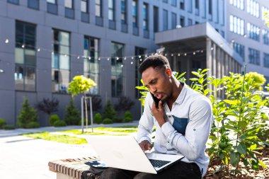 Afro-Amerikan erkek serbest çalışan uzaktan kumandalı borsa simsarı cep telefonu ve dizüstü bilgisayar tüccarı ile konuşuyor. Şehir parkında bir erkek, modern şehirli bir iş adamı.