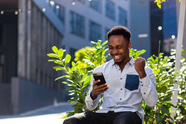 Mutlu Afro-Amerikan adam açık havada bankta oturuyor, elinde cep telefonuyla iyi haberler okuyor, dışarıda akıllı telefon sevinçlerini seyrederek zaferi kazanıyor. Erkek duyguları öğrenci sonuçları