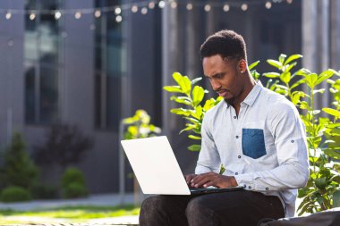 İnternet 'te çalışan siyahi serbest çalışan bir adam dizüstü bilgisayarını kullanarak şehir parkındaki modern ofis binasının önündeki bankta oturuyor. Açık havada klavyede yazı yazan mutlu Afrikalı Amerikalı öğrenci.