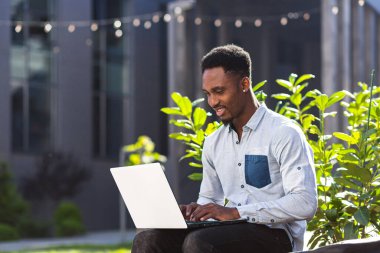 İnternet 'te çalışan siyahi serbest çalışan bir adam dizüstü bilgisayarını kullanarak şehir parkındaki modern ofis binasının önündeki bankta oturuyor. Açık havada klavyede yazı yazan mutlu Afrikalı Amerikalı öğrenci.