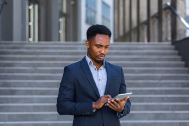Resmi takım elbiseli genç Afro-Amerikan iş adamı dışarıda arka plandaki modern ofis binasında elinde tabletle çalışıyor. Şehir dışında akıllı telefon ya da cep telefonu kullanan bir adam.