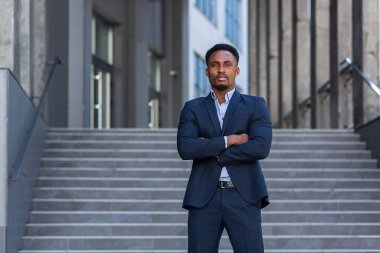 Genç özgüvenli Afro-Amerikan iş adamının modern ofis binasının arka planında duran ciddi iş adamına baktığı portre. Erkek, kollarını kavuşturmuş resmi bir takım elbise.