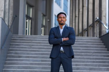 Genç özgüvenli Afro-Amerikan iş adamının modern ofis binasının arka planında duran ciddi iş adamına baktığı portre. Erkek, kollarını kavuşturmuş resmi bir takım elbise.