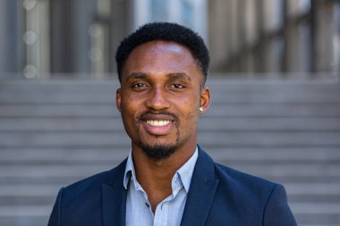 Afro-Amerikalı mutlu iş adamı dışarıda kameraya bakıyor. Gülümsemenin yüzü, modern şehir caddesinde resmi takım elbiseli yakışıklı siyah iş adamı kapalı arka planı bulanıklaştırdı.