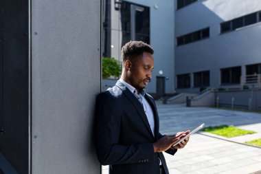 Resmi takım elbiseli genç Afro-Amerikan iş adamı dışarıda arka plandaki modern ofis binasında elinde tabletle çalışıyor. Şehir dışında akıllı telefon ya da cep telefonu kullanan bir adam.