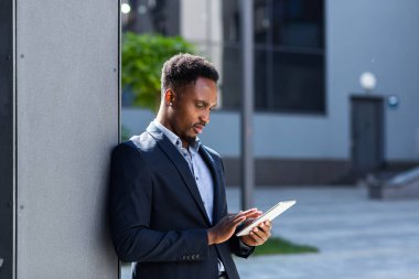 Resmi takım elbiseli genç Afro-Amerikan iş adamı dışarıda arka plandaki modern ofis binasında elinde tabletle çalışıyor. Şehir dışında akıllı telefon ya da cep telefonu kullanan bir adam.