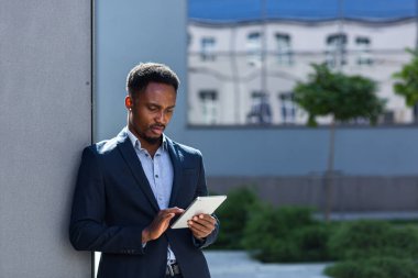 Resmi takım elbiseli genç Afro-Amerikan iş adamı dışarıda arka plandaki modern ofis binasında elinde tabletle çalışıyor. Şehir dışında akıllı telefon ya da cep telefonu kullanan bir adam.