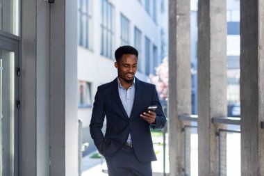 Başarılı genç, kendinden emin Afro-Amerikan iş adamı cep telefonuyla modern ofis arka planına karşı hareket ediyor. Siyah iş adamı resmi takım elbise içinde cep telefonuyla yürüyor.
