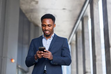 Başarılı genç, kendinden emin Afro-Amerikan iş adamı cep telefonuyla modern ofis arka planına karşı hareket ediyor. Siyah iş adamı resmi takım elbise içinde cep telefonuyla yürüyor.
